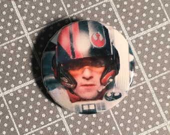 May include: A round button featuring a close-up image of a pilot wearing a helmet with a red stripe and a Rebel Alliance symbol. The pilot is looking directly at the viewer.