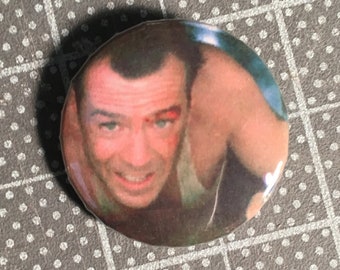 May include: A close-up of a round button featuring a photo of Bruce Willis as John McClane from the movie Die Hard. The button has a white border and a black background.
