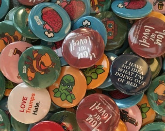 May include: A collection of colorful button pins with various designs and messages. Some of the pins feature humorous sayings, while others have illustrations of cartoon characters. The pins are a mix of red, blue, green, yellow, and white.