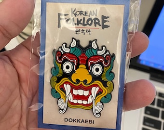 May include: A colorful enamel pin featuring a red, yellow, and blue Dokkaebi, a Korean folklore creature, with the text "Korean Folklore" and "Dokkaebi" in black.