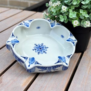 Może przedstawiać: Biała ceramiczna popielniczka z falistym brzegiem i niebieskimi wzorami kwiatowymi. W centrum znajduje się motyw niebieskiego kwiatu, a zewnętrzna krawędź ozdobiona jest niebieskimi liśćmi. Popielniczka posiada cztery wyraźne wgłębienia.