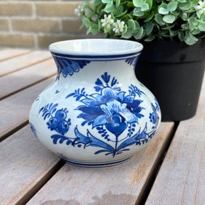 Może przedstawiać: Biały ceramiczny wazon o zaokrąglonym korpusie i rozszerzanym wylocie, ozdobiony ręcznie malowanymi niebieskimi wzorami kwiatów i liści. Powierzchnia posiada subtelne spękania szkliwa oraz niebieskie pasy wokół górnej i dolnej krawędzi naczynia.