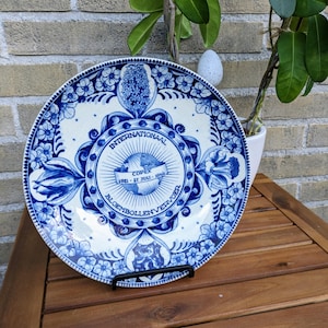 May include: A blue and white decorative plate with floral and geometric patterns. The plate features the text "INTERNATIONAAL BLOEMBOLLENVERVOER" and "COPEX 1981-21 JULI-1949". The plate is displayed on a black metal stand.