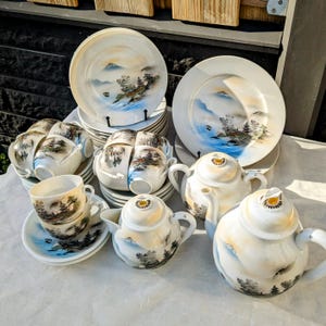 May include: A vintage Japanese tea set with a delicate floral design. The set includes a teapot, teacups, saucers, and a sugar bowl. The teacups and saucers feature a mountain scene with a blue and white color scheme.