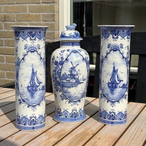 Pode incluir: Dois vasos cilíndricos altos e um pote com tampa com desenhos pintados à mão em azul e branco. Os vasos exibem veleiros, enquanto o pote retrata uma cena de moinho de vento. Cada peça é decorada com bordas e padrões florais detalhados.