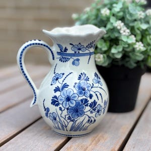 Pode incluir: Um jarro de cerâmica branca com um padrão floral azul pintado à mão. O jarro tem uma pega curva com um rebordo azul pontilhado, uma borda superior recortada e um corpo arredondado decorado com várias flores, folhas e caules azuis.