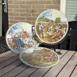 May include: Three circular glass plates featuring detailed illustrations of seasonal activities. One plate shows a winter scene with a castle, another depicts a summer harvest with workers, and the third shows spring planting. Each plate has a scroll label.