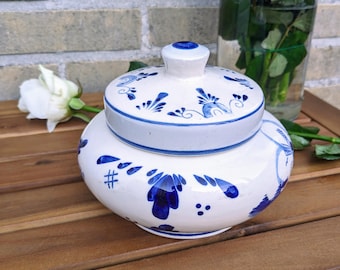 May include: A white ceramic jar with a lid, decorated with blue floral patterns. The jar has a rounded shape and a small knob on the lid. The design is reminiscent of Delftware. The jar is on a wooden surface.