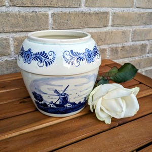 May include: A white ceramic pot with blue floral patterns and a windmill scene. The pot is round with a lid and sits on a wooden surface next to a white rose.
