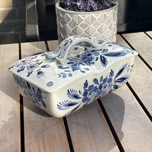 Könnte beinhalten: Eine rechteckige Butterdose aus Keramik mit einem Deckel, der einen geschwungenen Griff besitzt. Die cremeweiße Oberfläche ist mit handgemalten blauen Blumenmustern verziert, die verschiedene Blüten, Blätter und Ranken auf Deckel und Dose zeigen.