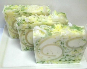 May include: Three handmade soap bars with a white base, featuring green and yellow accents, and a spiral design. The soaps are arranged on a white rectangular plate, creating a clean and natural aesthetic.