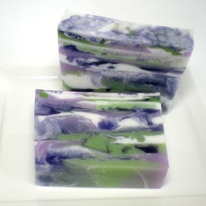 May include: Two rectangular bars of handmade soap with a layered design. The soap features swirls of white, green, purple, and dark blue. The bars are displayed on a white rectangular plate.