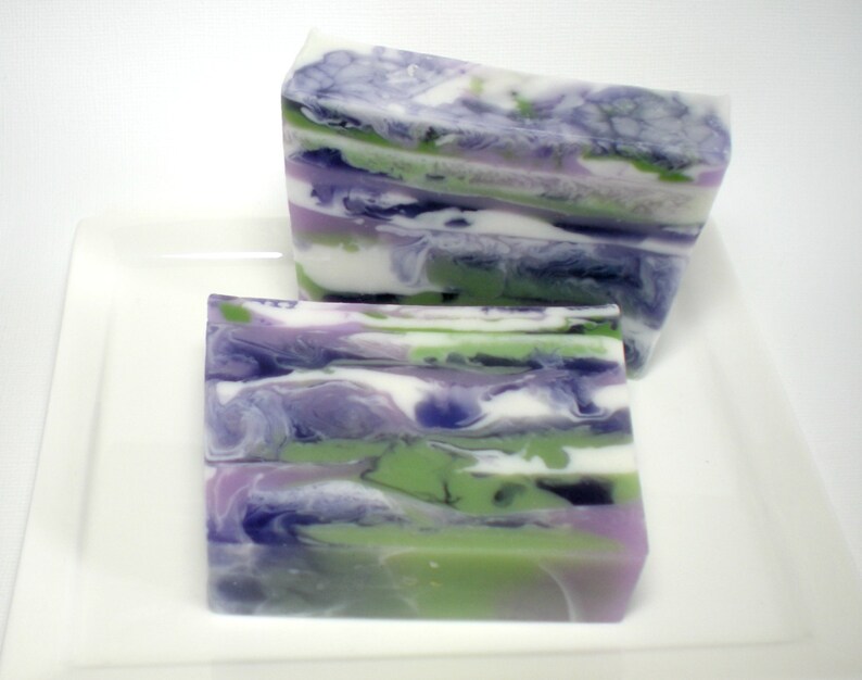 May include: Two rectangular bars of handmade soap with a layered design. The soap features swirls of white, green, purple, and dark blue. The bars are displayed on a white rectangular plate.
