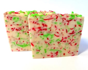 Op de afbeelding: Twee rechthoekige blokken gebroken witte zeep met een confetti-achtig patroon van kleine, verspreide rode en felgroene schilfers die in het oppervlak zijn verwerkt. De textuur oogt glad met zichtbare kleurspikkels over de blokken zeep.