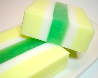 May include: Two bars of handmade soap with a layered design. The soap features stripes of yellow, white, and green. The bars are rectangular and sit on a white surface. The soap appears to be freshly made.