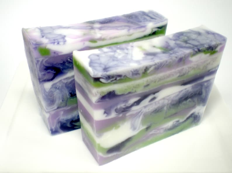May include: Two rectangular bars of handmade soap with a layered design. The soap features swirls of purple, green, white, and black, creating a marbled effect. The bars are stacked on a white surface.