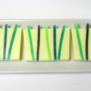 Op de afbeelding: Drie handgemaakte zeepblokken met een lichtgele basis en verticale strepen in groen- en zwarttinten. De zepen zijn gerangschikt op een wit rechthoekig bord, met een modern, minimalistisch ontwerp.