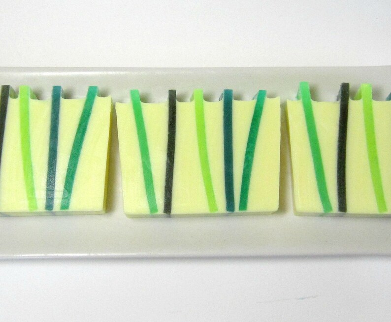 Op de afbeelding: Drie handgemaakte zeepblokken met een lichtgele basis en verticale strepen in groen- en zwarttinten. De zepen zijn gerangschikt op een wit rechthoekig bord, met een modern, minimalistisch ontwerp.
