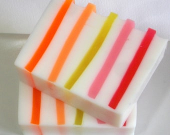 Op de afbeelding: Twee handgemaakte zeepblokken met een witte basis en kleurrijke strepen in oranje, geel, roze en rood. De blokken zijn diagonaal gestapeld, wat het levendige ontwerp laat zien. De zeep heeft een strakke, frisse uitstraling.