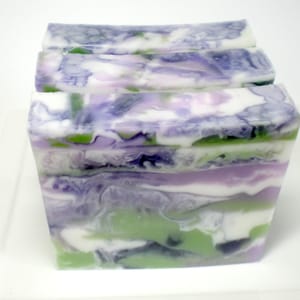 May include: Four rectangular bars of handmade soap with a marbled design. The soap features layers of white, lavender, green, and dark purple colors. The bars are stacked on top of each other, creating a visually appealing pattern.