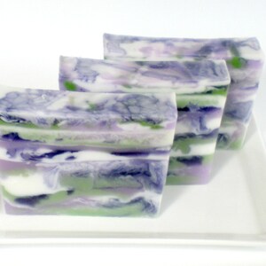May include: Three rectangular bars of handmade soap with a layered design. The soap features swirls of white, green, purple, and blue colors. The bars are arranged on a white rectangular plate.