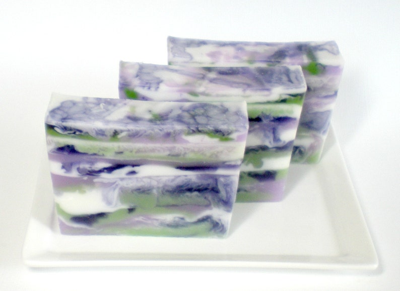 May include: Three rectangular bars of handmade soap with a layered design. The soap features swirls of white, green, purple, and blue colors. The bars are arranged on a white rectangular plate.