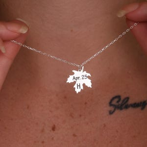 May include: A silver necklace featuring a maple leaf pendant. The pendant is engraved with "Apr. 25" and the letter "H". The necklace is held by a person.
