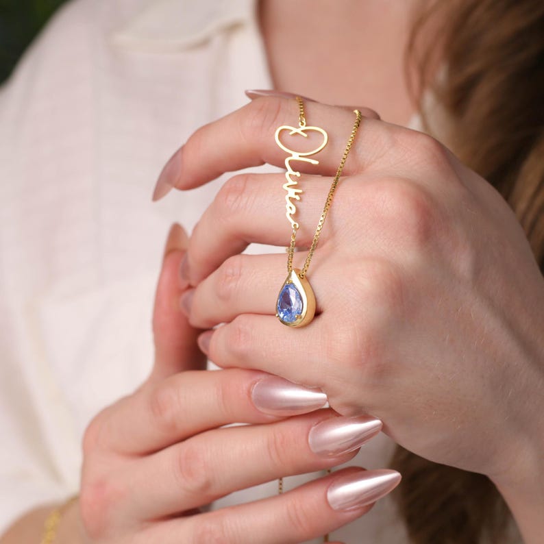 K&ouml;nnte beinhalten: Goldkette mit einem tropfenf&ouml;rmigen blauen Edelstein-Anh&auml;nger und einem personalisierten Namensschild mit der Aufschrift "Olivia".