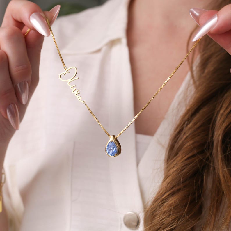 K&ouml;nnte beinhalten: Eine goldene Halskette mit einem tropfenf&ouml;rmigen Anh&auml;nger, der mit einem blauen Edelstein besetzt ist. Die Halskette hat au&szlig;erdem ein zartes Namensschild mit dem Namen "Olivia" in Kursivschrift.