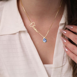 K&ouml;nnte beinhalten: Eine goldene Halskette mit einem tropfenf&ouml;rmigen blauen Edelstein-Anh&auml;nger und einem Anh&auml;nger mit dem Namen "Olivia" in Schreibschrift.