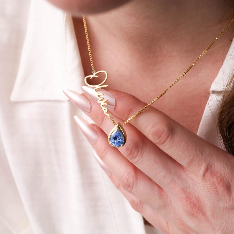 K&ouml;nnte beinhalten: Eine goldene Halskette mit einem tropfenf&ouml;rmigen blauen Edelstein-Anh&auml;nger und einem Anh&auml;nger mit einem kursiv geschriebenen Namen, der "Olivia" lautet.