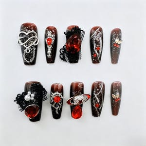 Op de afbeelding: Een set van tien kunstnagels met een donkerrode en bruine gradiënt. Elke nagel heeft unieke versieringen, waaronder zilveren bedels, rode edelstenen en zwarte kanten accenten. Sommige nagels hebben een glitterafwerking.
