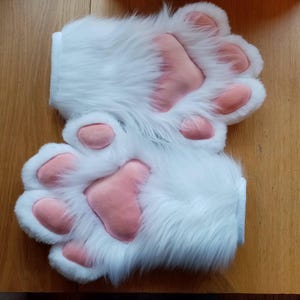 May include: A pair of white furry paw gloves with pink paw pads. The gloves are made of soft, plush fabric and have a realistic paw design.