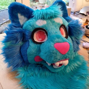 Op de afbeelding: Een blauwe en roze harige dierenkop met een tandenrij. De kop is gemaakt van pluche stof en heeft grote, ronde ogen. De kop is een kostuumstuk voor cosplay of furry fandom.