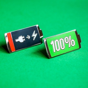 Può includere: Due spille di metallo a forma di batteria. Una spilla è nera con una striscia rossa e una spina bianca e un fulmine. L'altra spilla è verde con il testo "100%" in bianco.