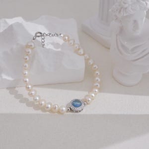 Op de afbeelding: Een delicate armband met crèmewitte parels. De armband heeft een zilveren sluiting en een centrale bedel met een lichtblauwe steen, omringd door kleine, heldere stenen. De sieraden worden gepresenteerd op een wit oppervlak.