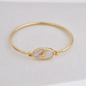 Puede incluir: Brazalete dorado con un centro ovalado de nácar. El centro presenta un diseño de estrella de cuatro puntas con un pequeño acento redondo. El brazalete tiene un cierre sencillo.