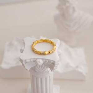 May include: A gold-colored ring with a bamboo design, adorned with small, sparkling stones. The ring is displayed on a white pedestal, with a blurred white background. The ring's design features textured segments and is a piece of jewelry.