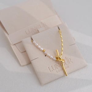 Può includere: Una collana d'oro con un ciondolo annodato e una fila di perle bianche. La collana è esposta su un sacchetto di velluto bianco con il nome del marchio "Lungur London" stampato su di esso.