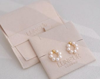 May include: A pair of pearl hoop earrings with gold accents, displayed on a beige card with the brand name "LUNEUR LONDON" embossed. A matching beige pouch is visible in the background. The earrings are a classic jewelry item.