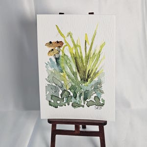 May include: Watercolor painting of green and yellow plants and brown mushrooms on white paper. The artwork is displayed on a small wooden easel. The artist's initials, SEB, are visible in the lower right corner.