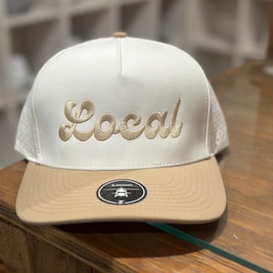 May include: A white and tan baseball cap with a mesh back. The front of the cap has the word "Local" embroidered in gold.