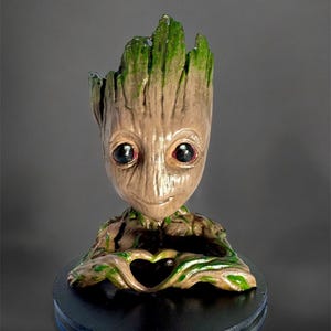 May include: A brown and green figurine of a character from the Guardians of the Galaxy movie series. The character is making a heart shape with its hands. The figurine is on a black base.