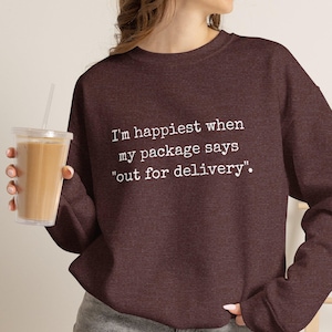 May include: A dark maroon long-sleeve sweatshirt featuring white text that reads, I'm happiest when my package says "out for delivery". The garment has a crew neckline and is paired with gray denim shorts, while a clear plastic cup is held.