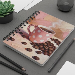 Può includere: Quaderno a spirale con una tazza di caffè rosa a pois e vapore, circondata da chicchi di caffè. L'opera d'arte utilizza una calda tavolozza di rosa, marroni e creme, evocando un'atmosfera accogliente da caffetteria.