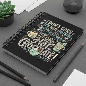 Può includere: Quaderno nero a spirale con la scritta "I don't care if it's hot outside, any day is a good day for hot chocolate!" in un carattere stravagante. La copertina presenta illustrazioni di tazze e cacao.