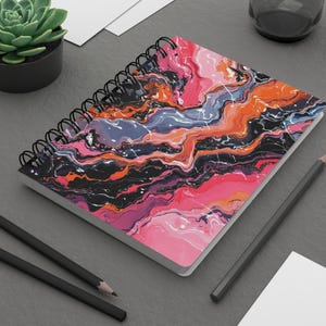 Può includere: Quaderno a spirale con un vivace design astratto. La copertina presenta un motivo a vortice di tonalità rosa, arancione, nero e blu con accenti bianchi. Sono visibili anche due matite nere e una piccola pianta succulenta.