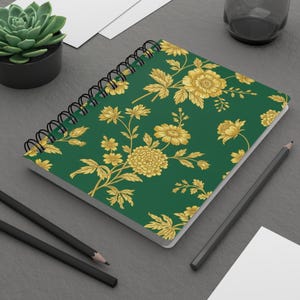 Può includere: Un taccuino a spirale con una copertina verde con un motivo floreale dorato. Il taccuino è aperto, rivelando pagine bianche. Sono visibili anche due matite nere e una piccola pianta grassa in un vaso nero.