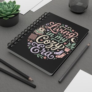Può includere: Quaderno nero a spirale con la scritta "Living in my Cozy Era" in un design colorato e stravagante. Il design include una tazza di caffè, libri, una sciarpa lavorata a maglia, una pianta in vaso e un calzino accogliente. Una pianta succulenta in un vaso nero è sullo sfondo.