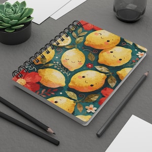 Può includere: Quaderno a spirale con un allegro motivo a limoni con facce sorridenti, fiori rossi e foglie verdi su sfondo verde acqua. Il quaderno ha una rilegatura a spirale nera. Sono visibili anche matite e una pianta succulenta.